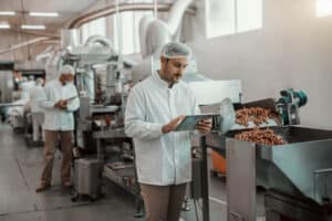 young caucasian serious supervisor evaluating quality food food plant while holding tablet man is dressed white uniform having hair net